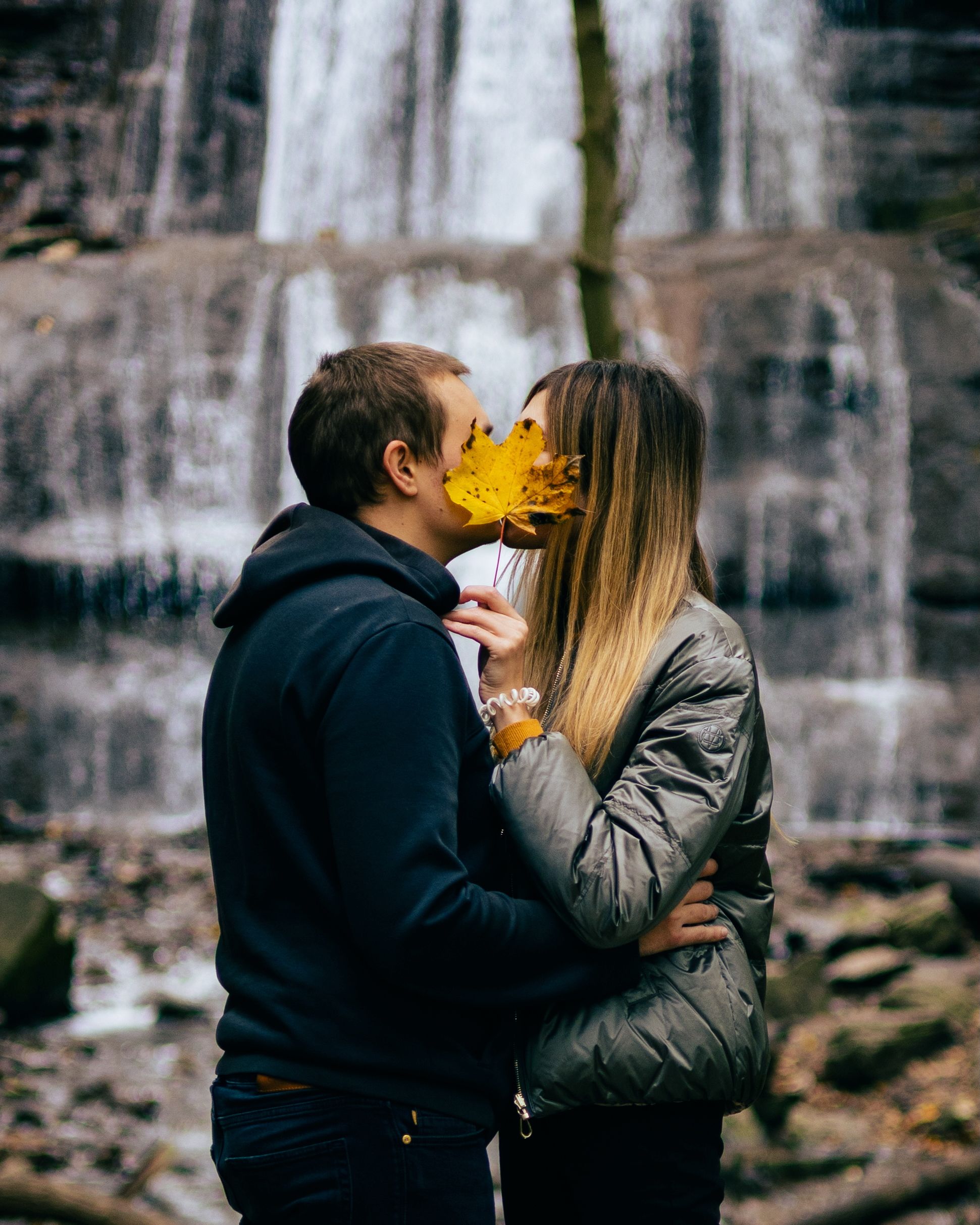 A couple kissing in front of a waterfall with an autumnal yellow leaf in front of their faces