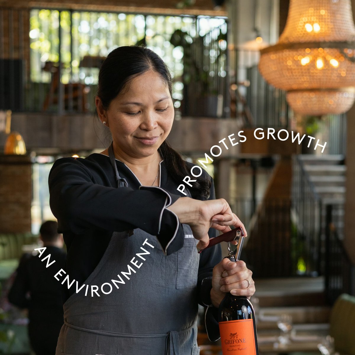 A server opening a bottle of wine using a corkscrew inside a restaurant, with overlaid text reading “AN ENVIRONMENT THAT PROMOTES GROWTH”.