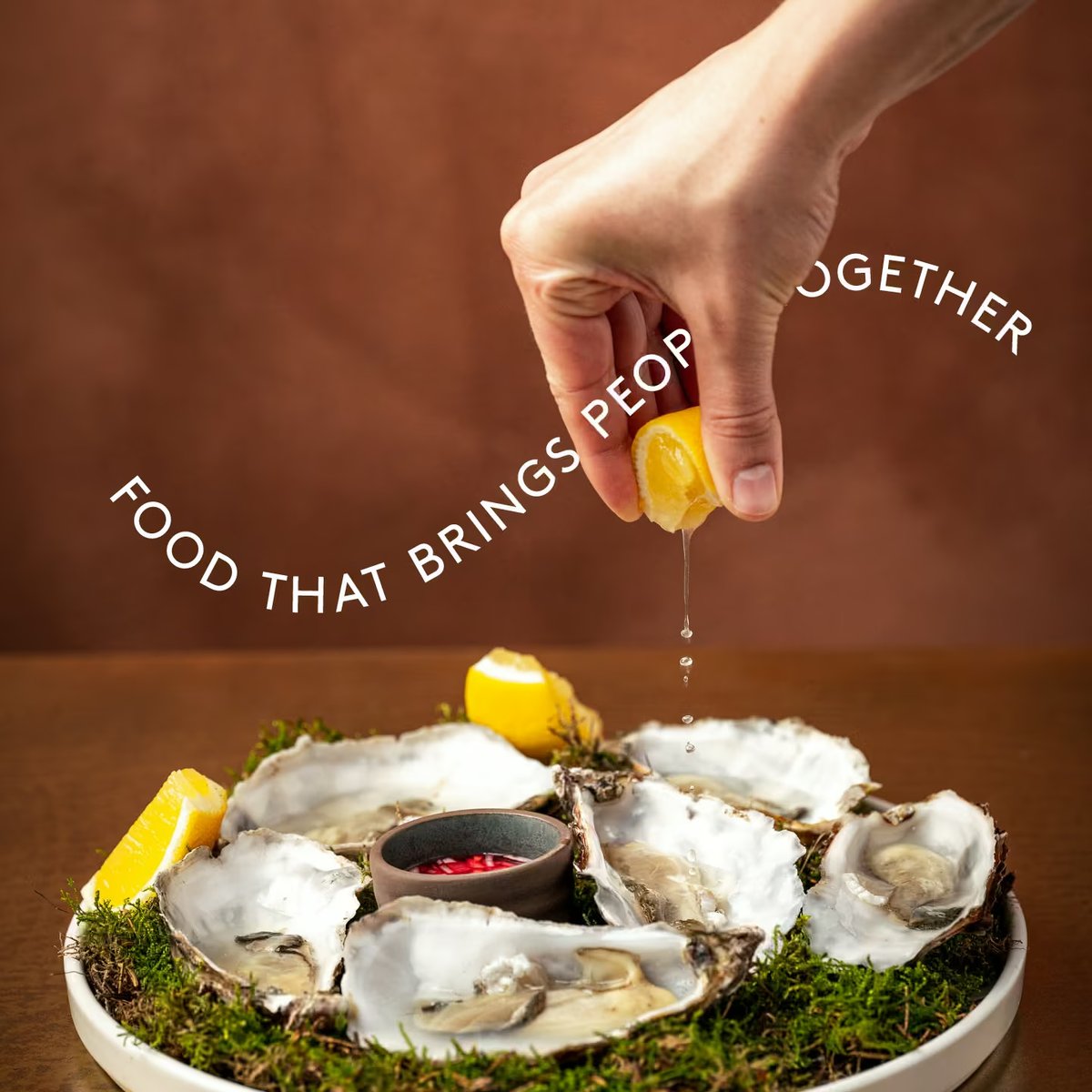 Hand squeezing fresh lemon juice over a platter of fresh oysters on the half shell with mignonette sauce. Overlay text reads “FOOD THAT BRINGS PEOPLE TOGETHER”.