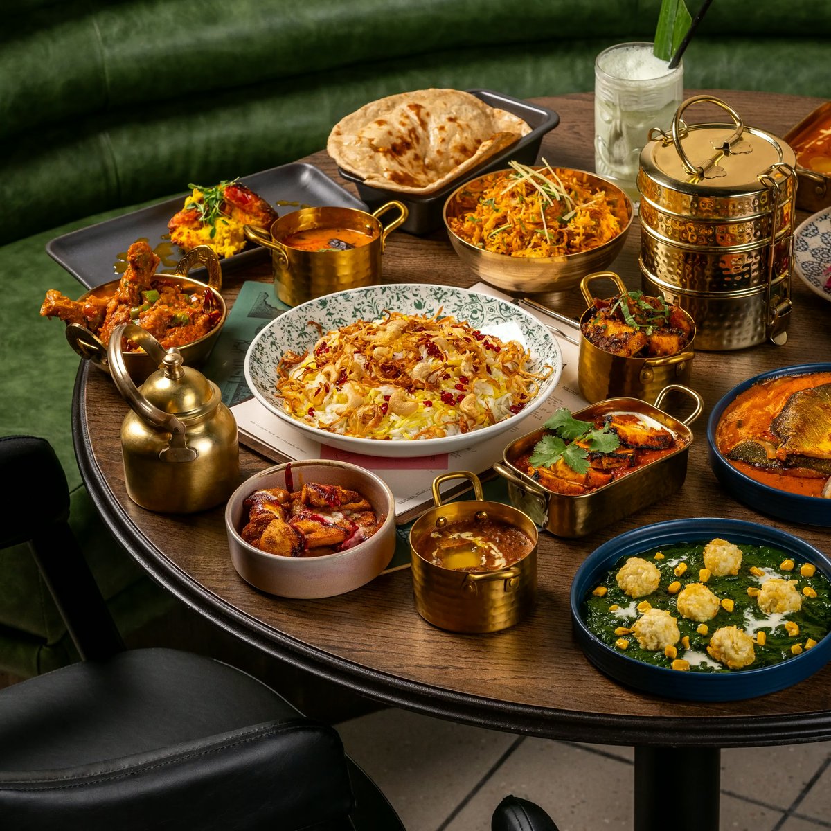 A round wooden dining table filled with a rich spread of Indian dishes served in traditional brass metal bowls, patterned ceramic plates, and a multi-tiered brass tiffin stack.