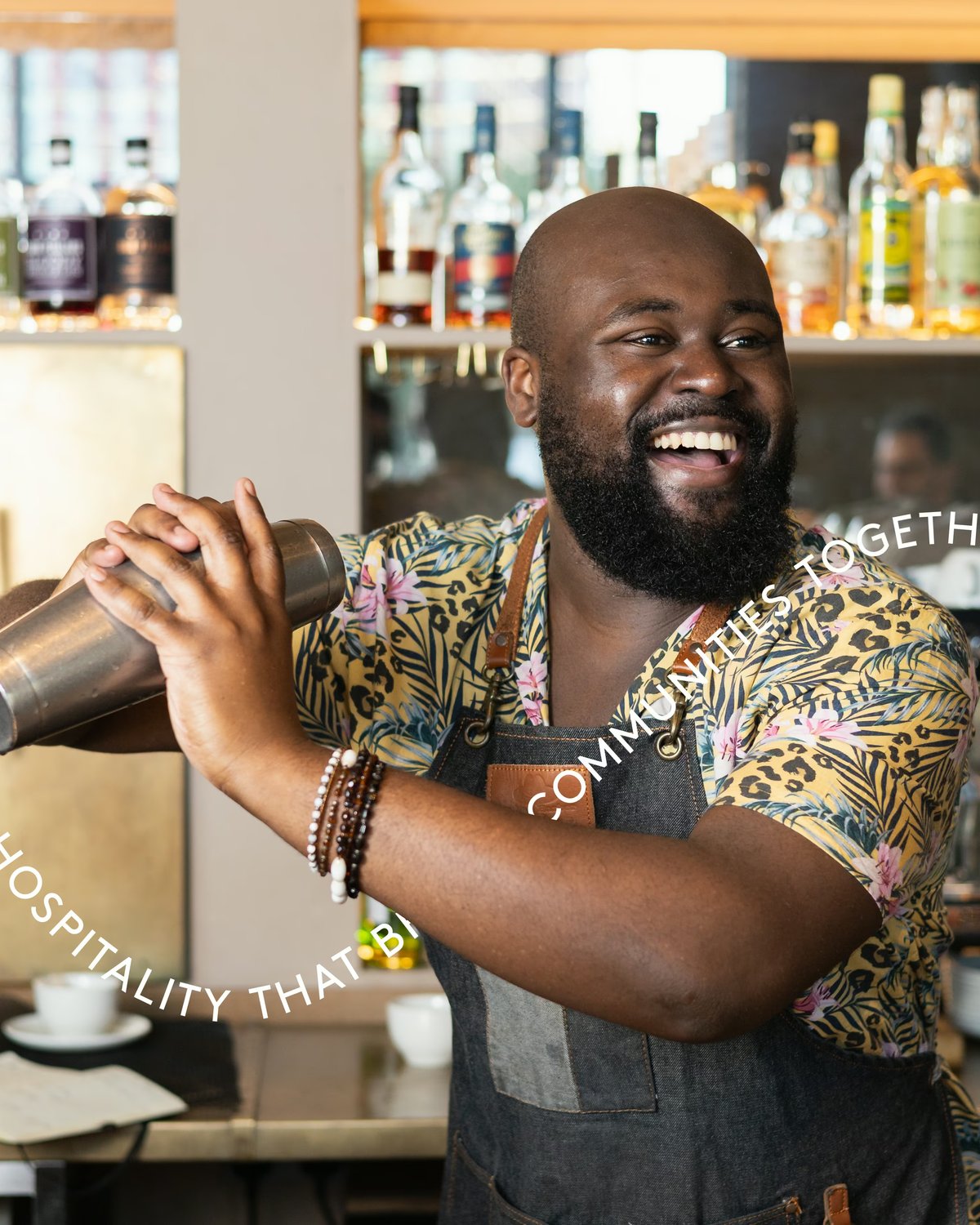 A smiling bartender shaking a cocktail in a metal shaker behind a bar, overlaid with curved text reading "Hospitality that brings communities together."