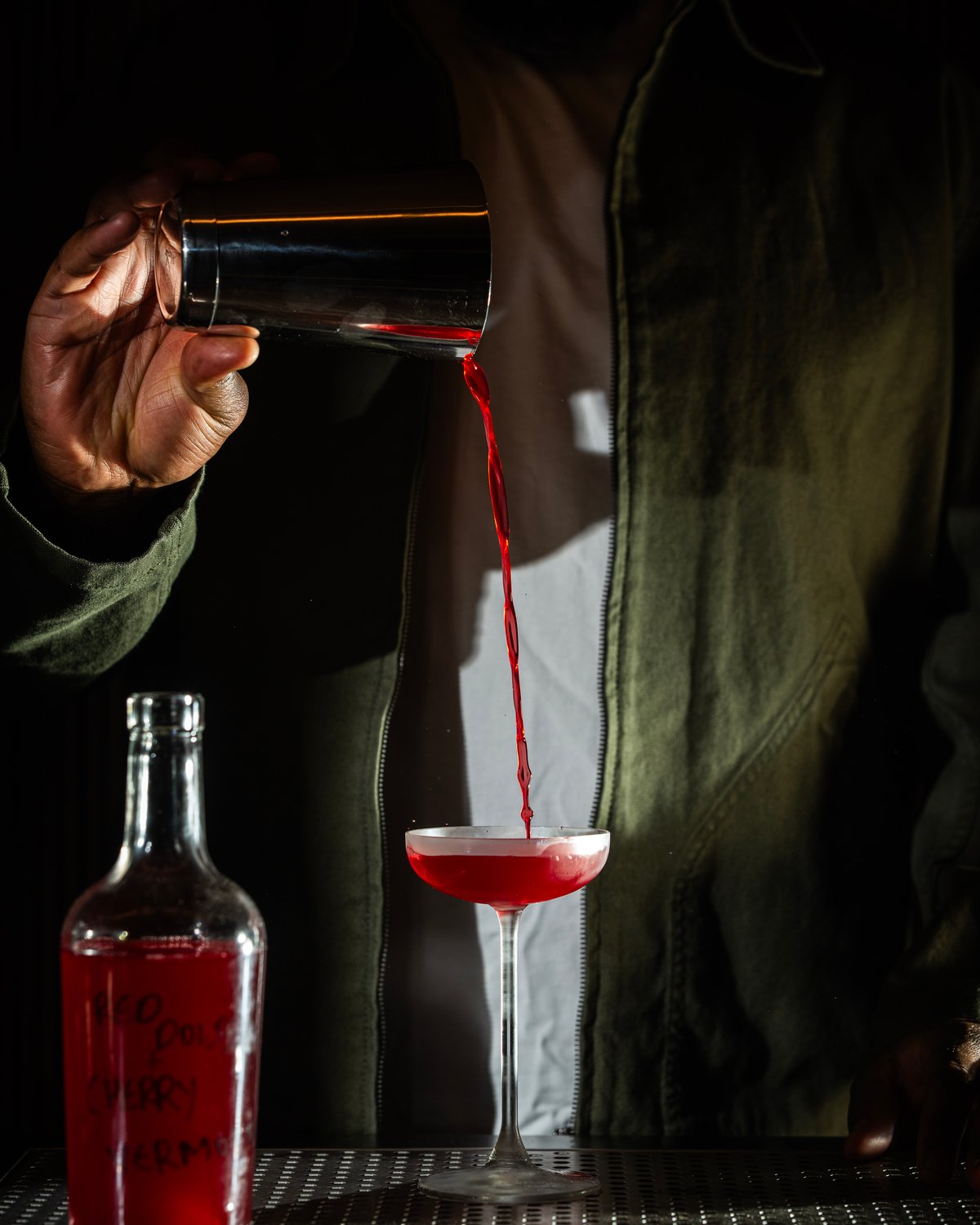 Moody, dramatic shot of a bartender pouring a bright red cocktail from a metal shaker into a coupe glass, next to a clear bottle labeled with handwritten red text on a bar counter.