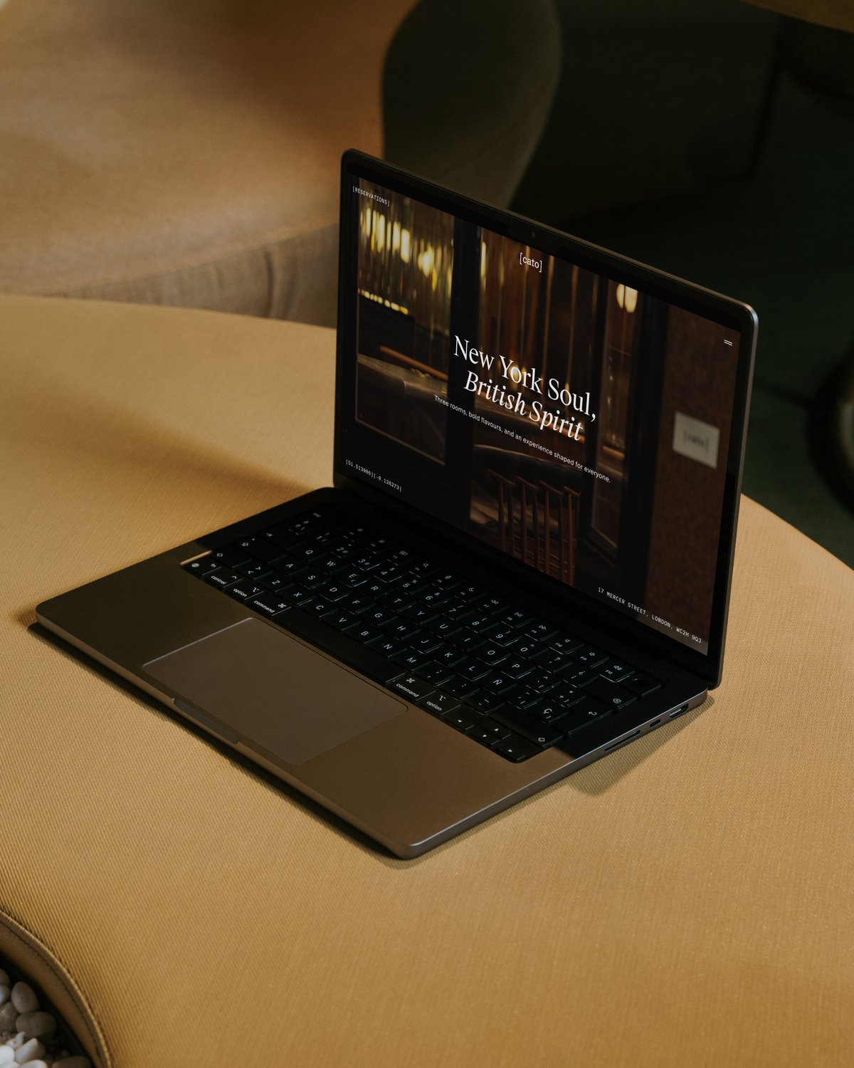 Angle view of an open laptop sitting on a curved mustard-yellow upholstered bench, displaying the Cato website homepage with the tagline "New York Soul, British Spirit".