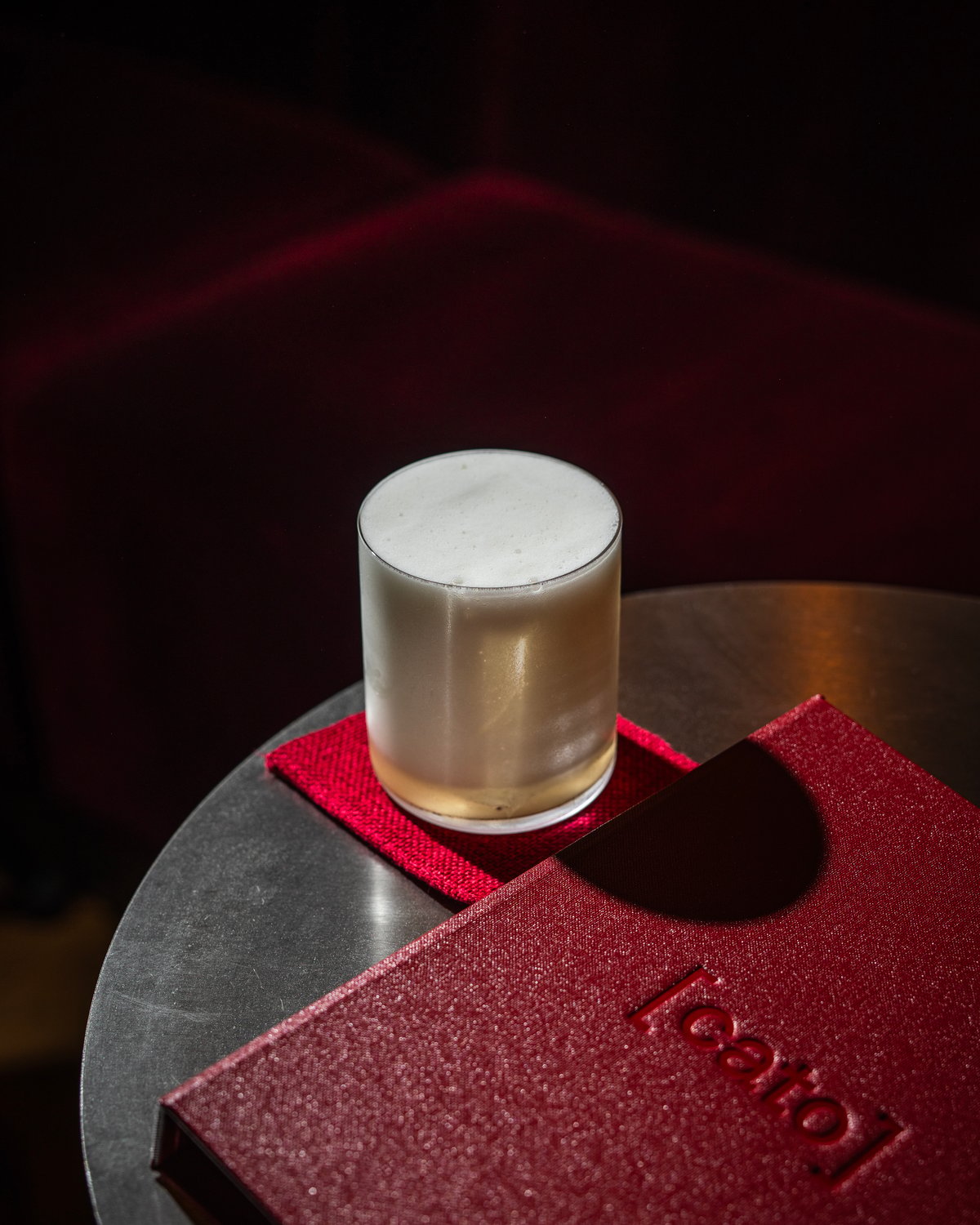 A foamy cocktail in a rocks glass sitting on a red coaster alongside a red, textured Cato menu on a round table.