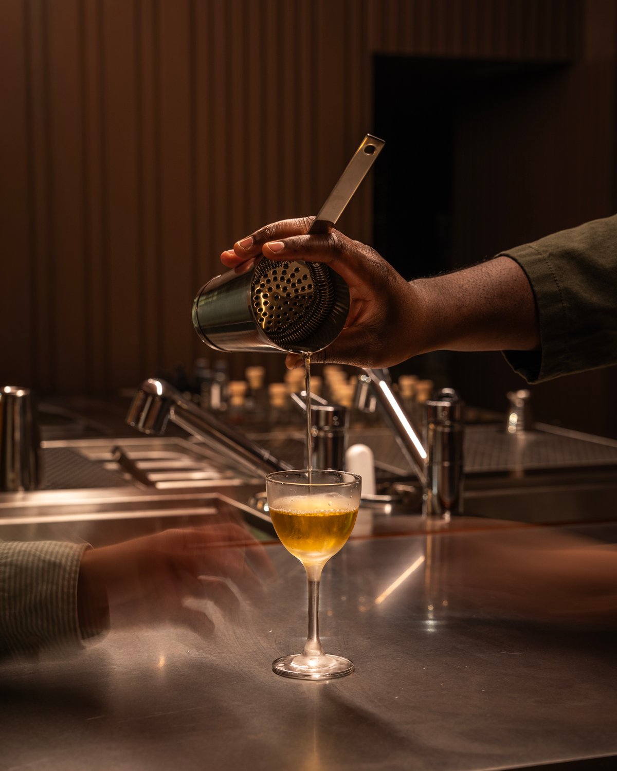 Close-up of a bartender pouring a golden cocktail from a metal shaker with a Hawthorne strainer into a stemmed glass at a dimly lit bar.