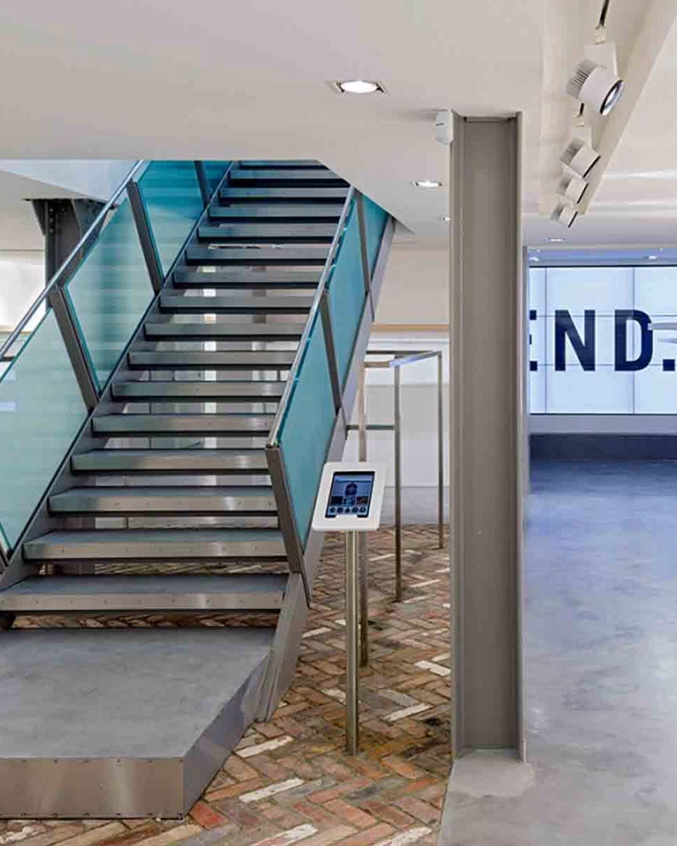 Modern retail store interior featuring a central metal staircase with frosted blue glass railings and a tablet display stand.