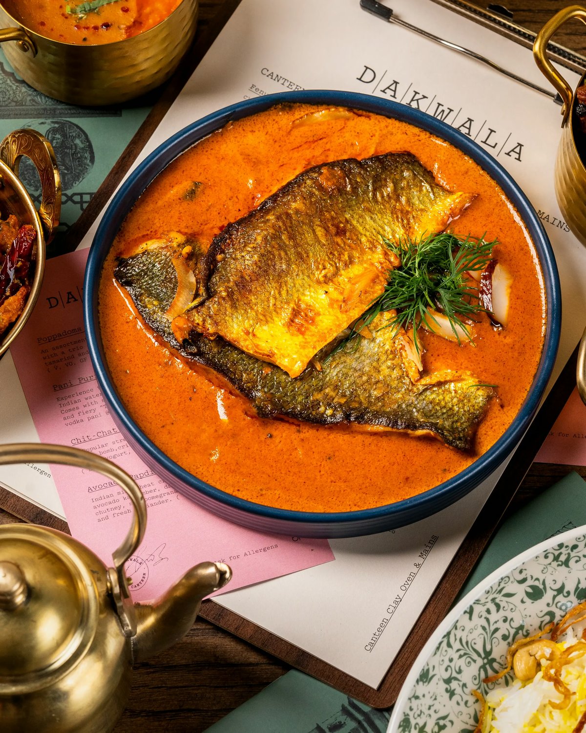 Close-up top-down shot of a blue bowl filled with orange Indian fish curry, topped with fresh herbs, resting on a Dakwala paper menu alongside small brass serving pots.