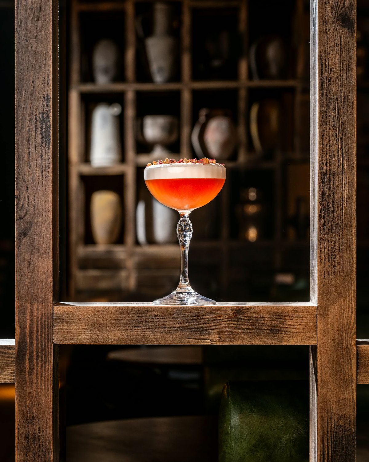 An orange-pink cocktail with a layer of white foam and rose petal garnish, served in an elegant Coupe glass framed by wooden shelving.