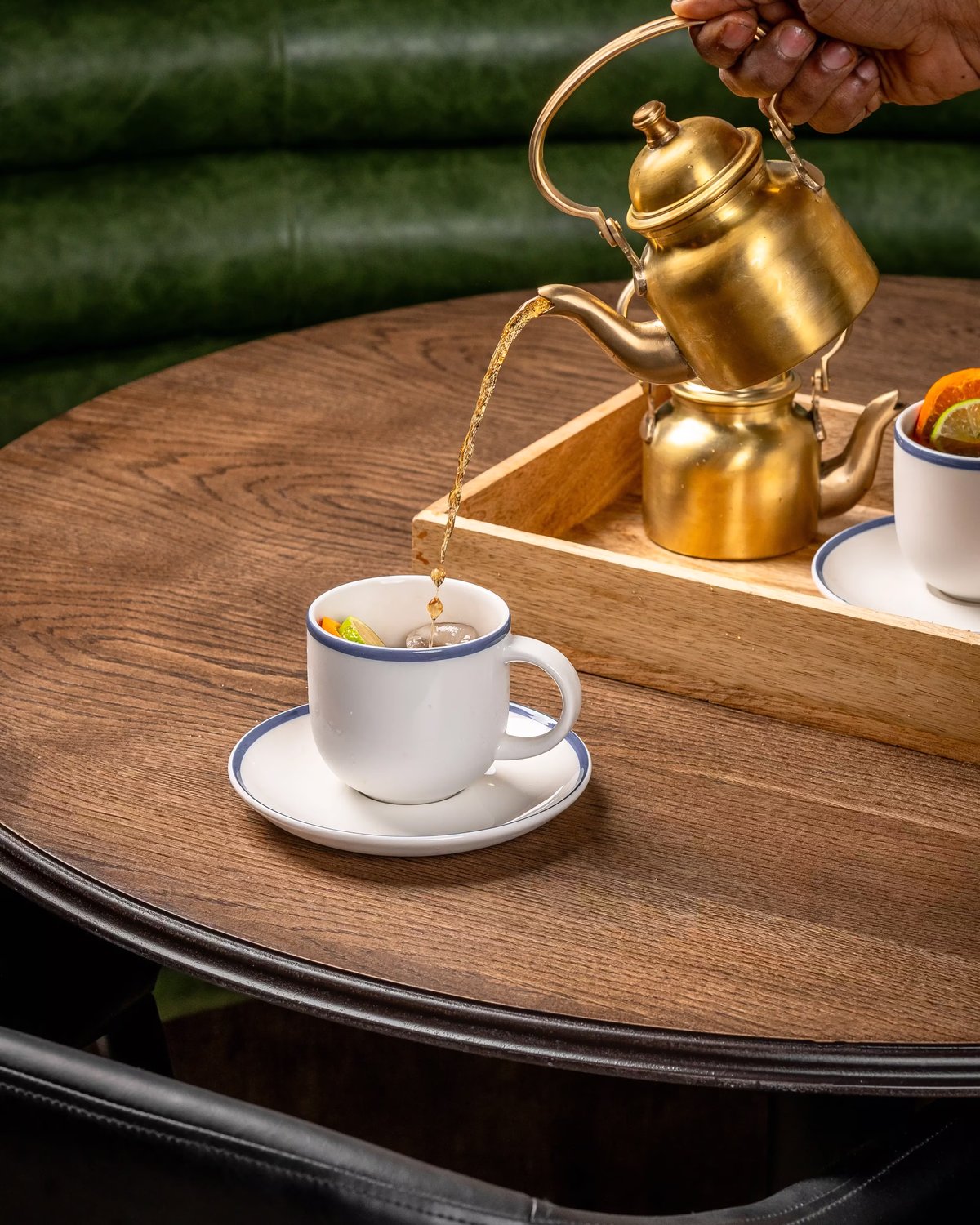 A person pouring a cocktail from an ornate brass kettle into a white porcelain cup with a blue trim, set on a dark wood table.