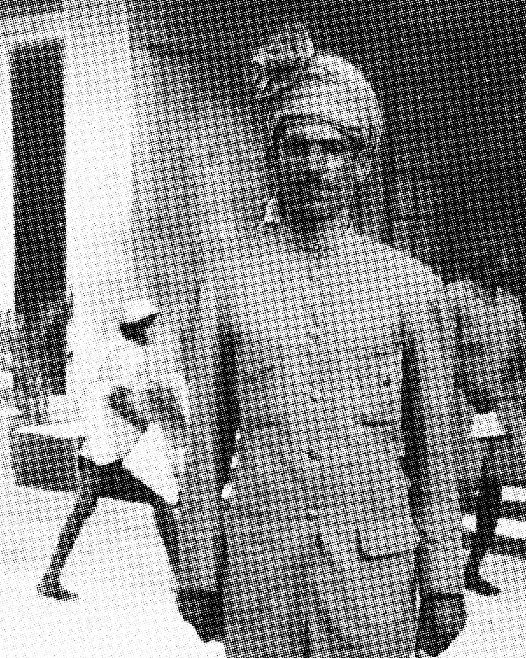 Black and white halftone archival photograph of a man in a formal buttoned tunic and traditional turban standing outdoors in front of a building.