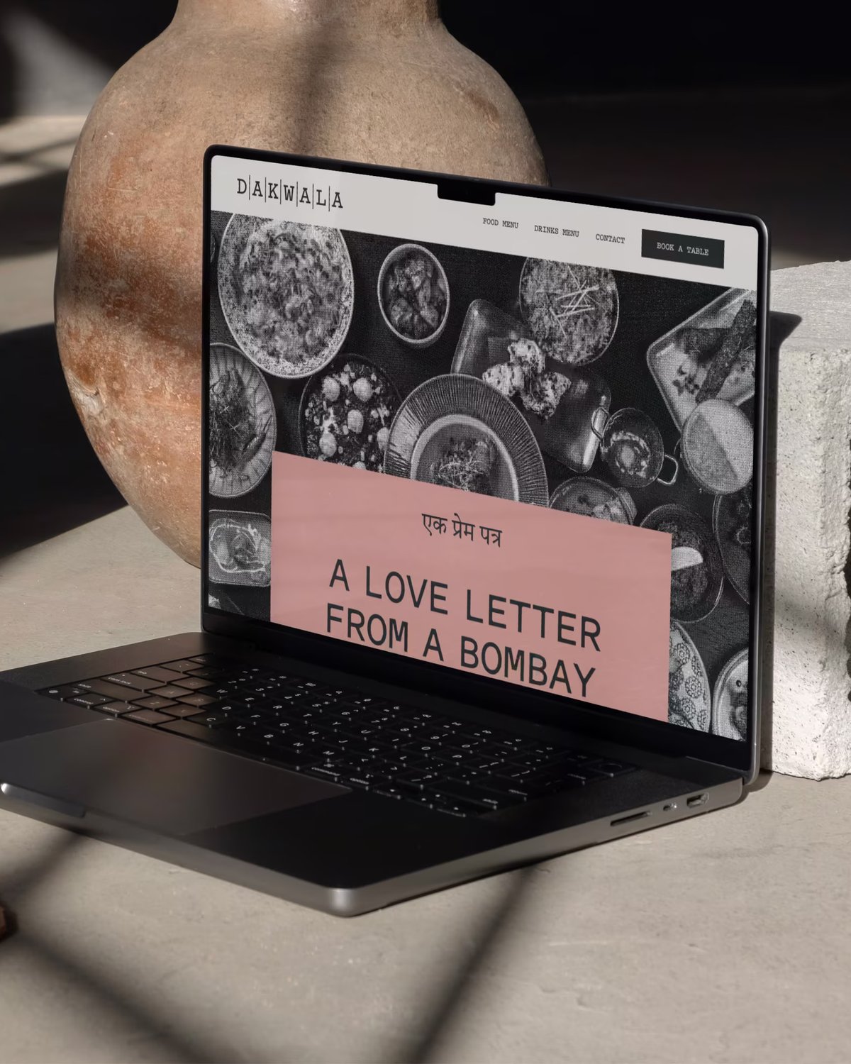 A dark laptop open on a concrete surface next to clay pottery and a cinder block, displaying the Dakwala website with the headline "A LOVE LETTER FROM A BOMBAY".