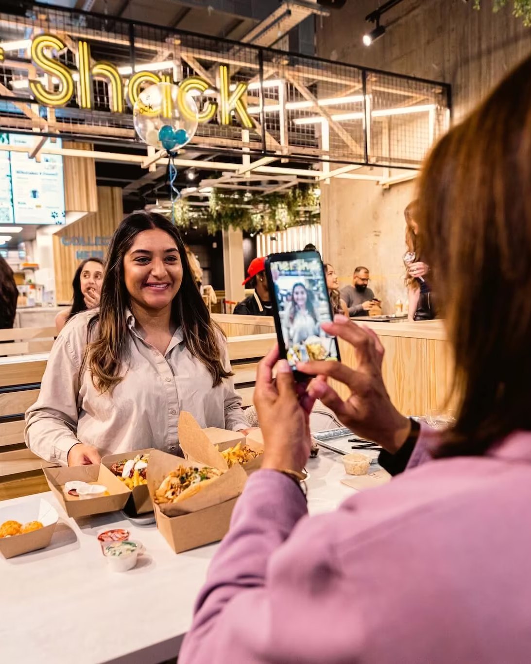 A woman sitting at a booth in a busy Doner Shack restaurant smiling while a friend takes her photo with a smartphone.