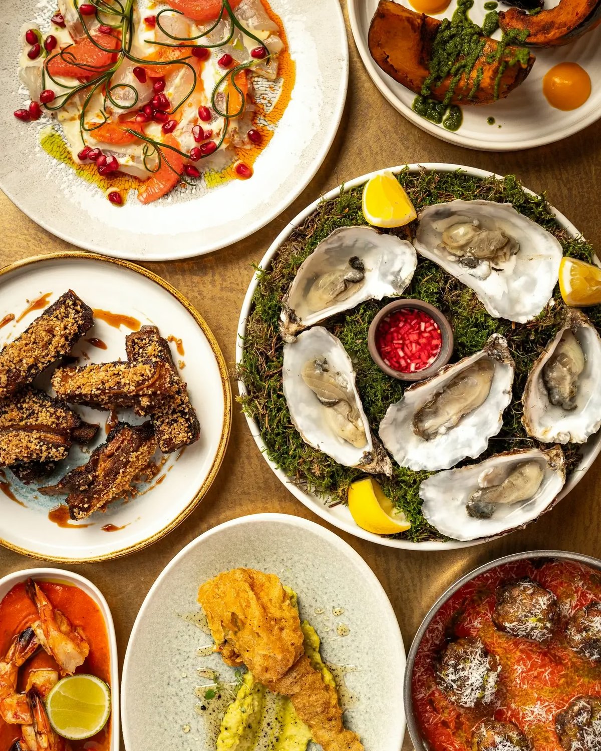 Top-down view of a varied dining spread featuring a central platter of fresh oysters, crudo, ribs, meatballs in red sauce, and roasted squash.