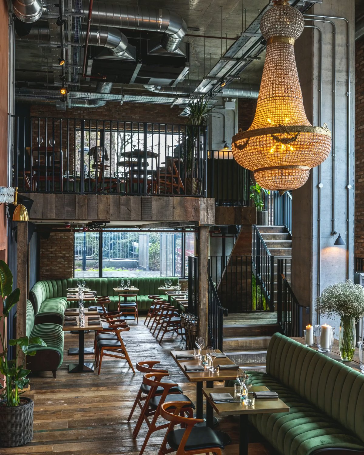 Modern restaurant interior at Fern featuring green velvet plush seating, wooden floors, exposed ceiling pipes, and a large crystal chandelier.