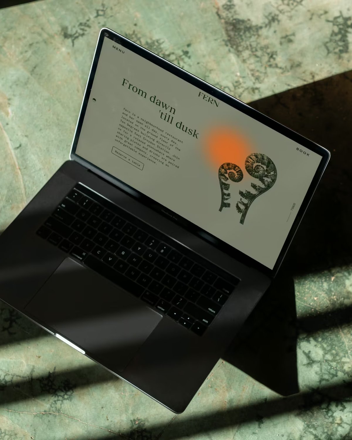 Top-down view of an open laptop sitting on a green textured floor, displaying the website homepage for Fern restaurant.