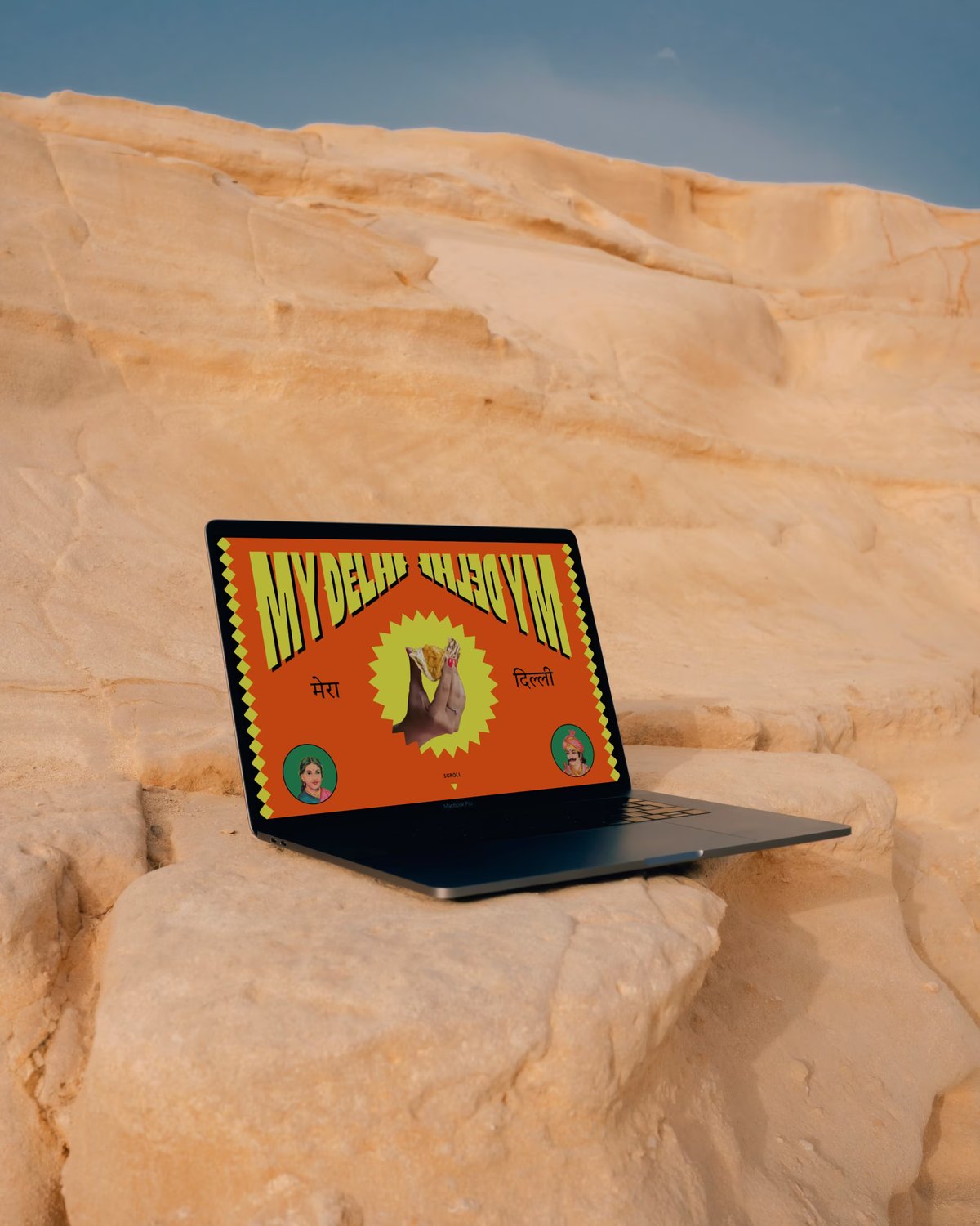 An open laptop sitting on a light-colored sandstone rock formation outdoors, displaying a colorful orange graphic with yellow text reading "MY DELHI" on the screen.