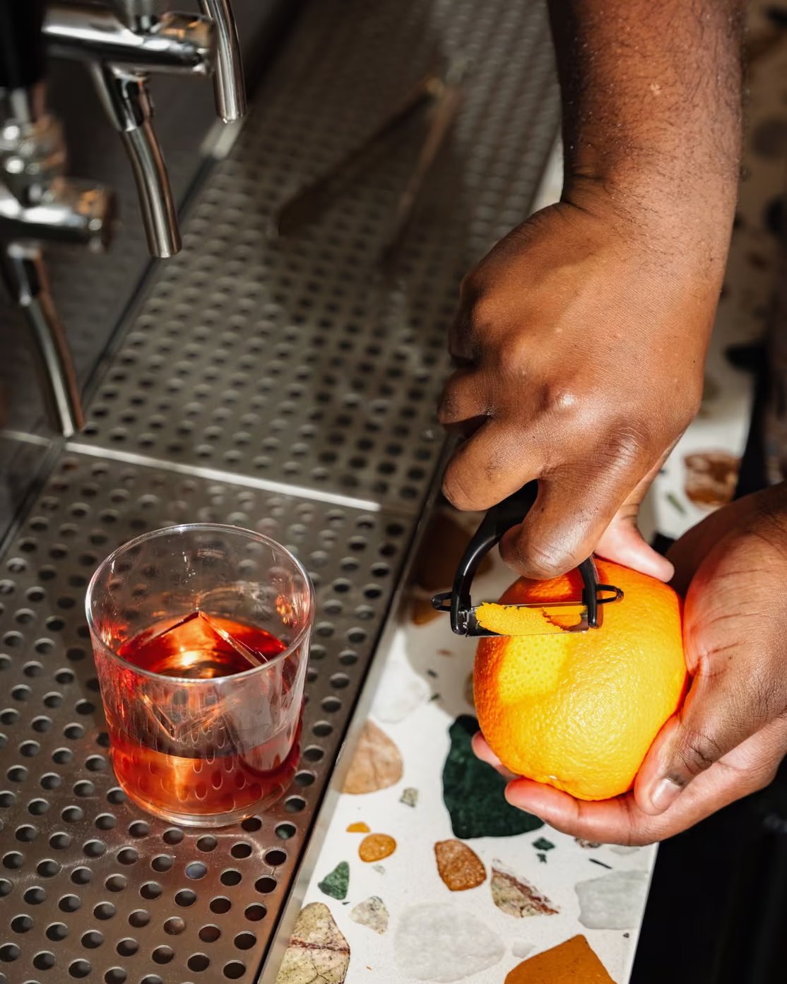 A bartender peeling an orange with a hand peeler beside a negroni with a large ice cube on a stainless steel drip tray.