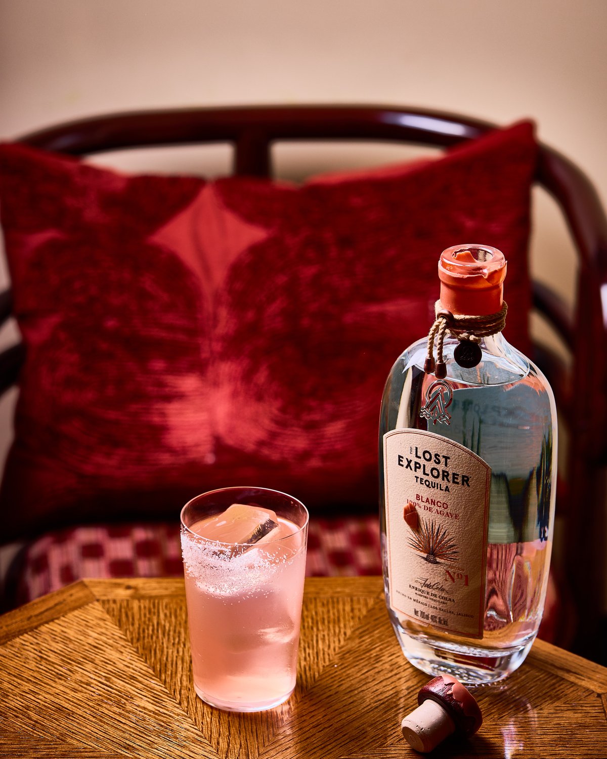 A tall salt-rimmed glass with a light pink cocktail served on ice next to a uncorked bottle of The Lost Explorer Tequila on a wooden table, with a red velvet chair in the background.