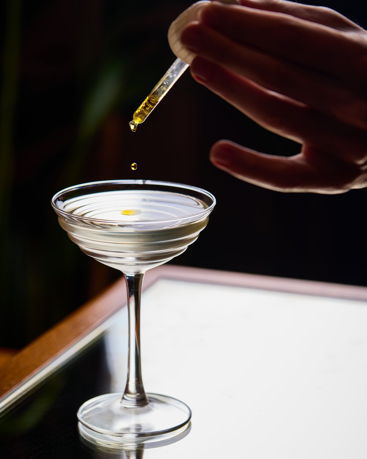 Close-up action shot of a hand holding a glass pipette dropper, releasing golden drops into a clear cocktail inside a ribbed coupe glass on a reflective tabletop.