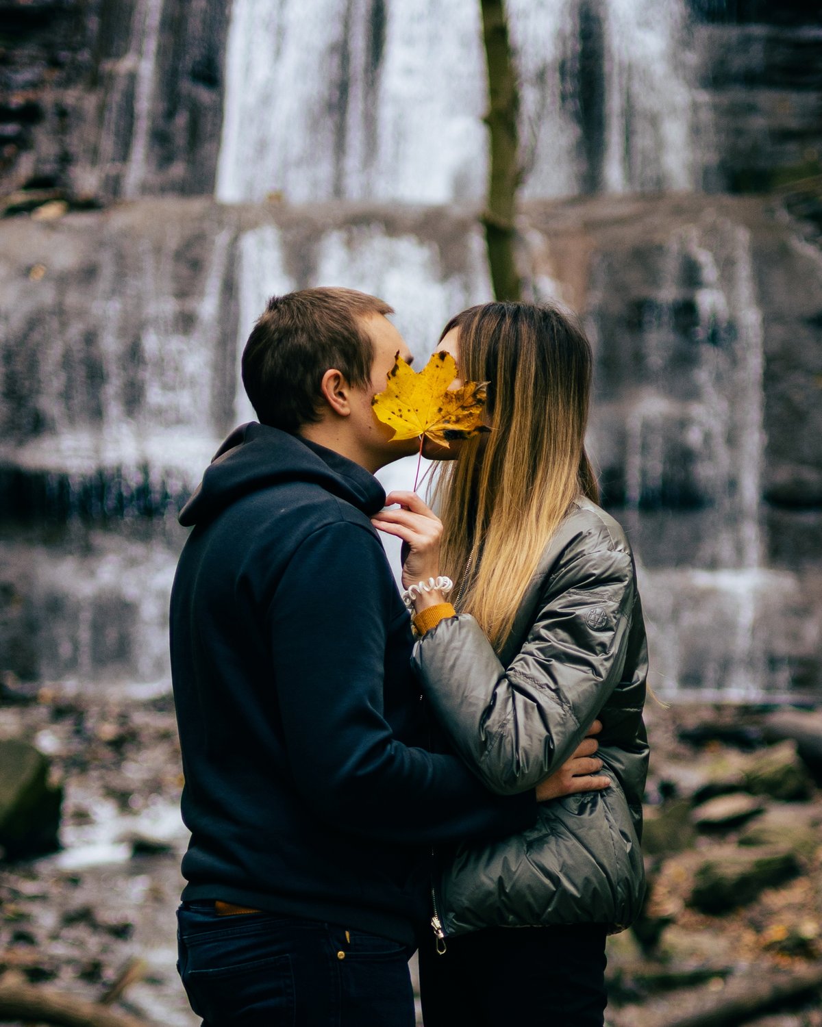 A couple kissing in front of a warefall with an autumnal yellow leaf in front of their faces