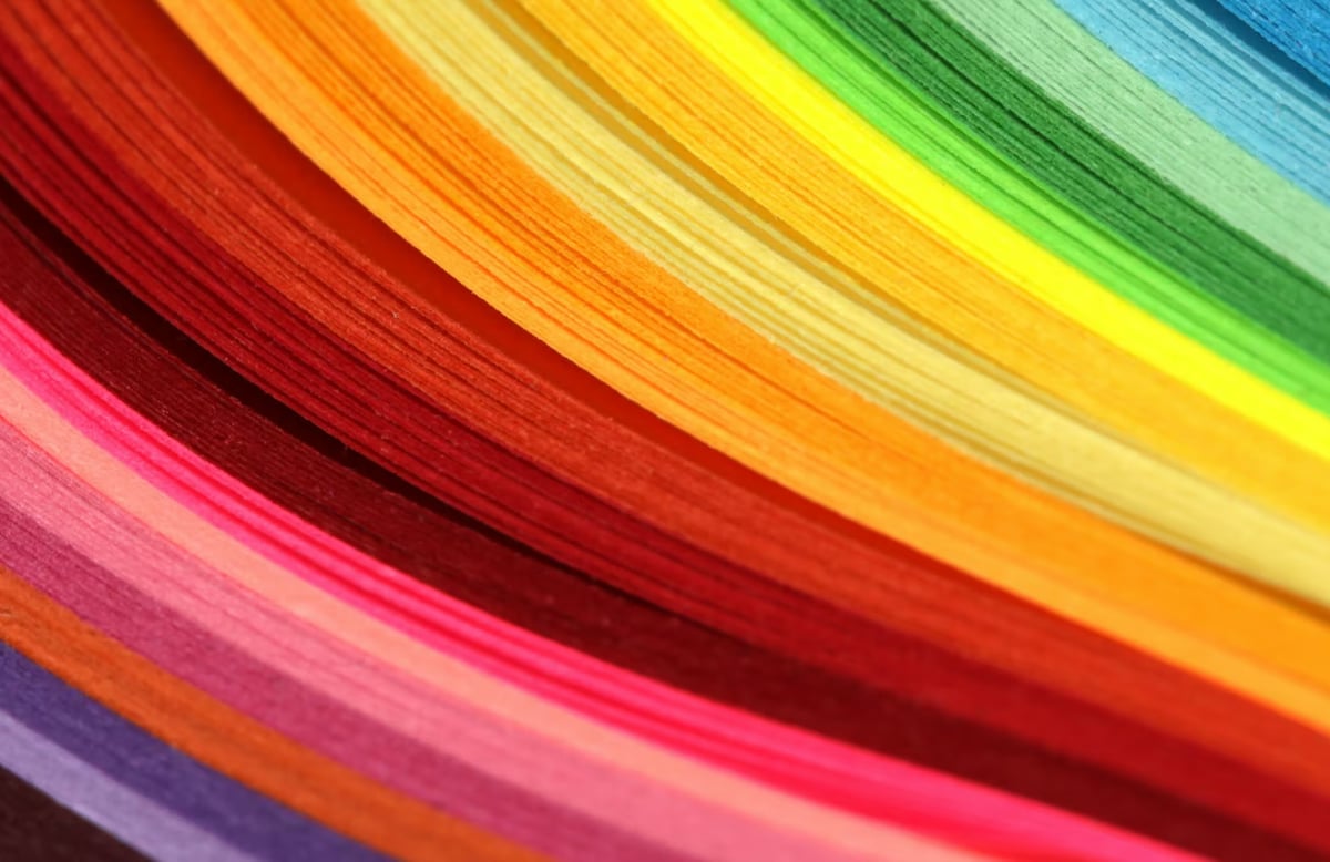 A close-up shot of thin, curved paper layers fanning out in a smooth color spectrum, transitioning from deep purple and pink into bright red, orange, yellow, green, and blue.