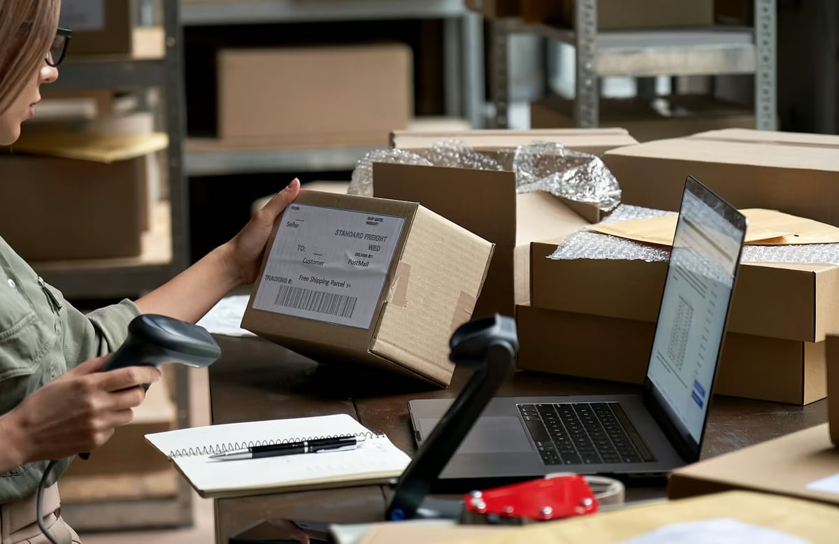A person in a warehouse scanning a shipping label on a cardboard box next to a laptop and packing supplies.
