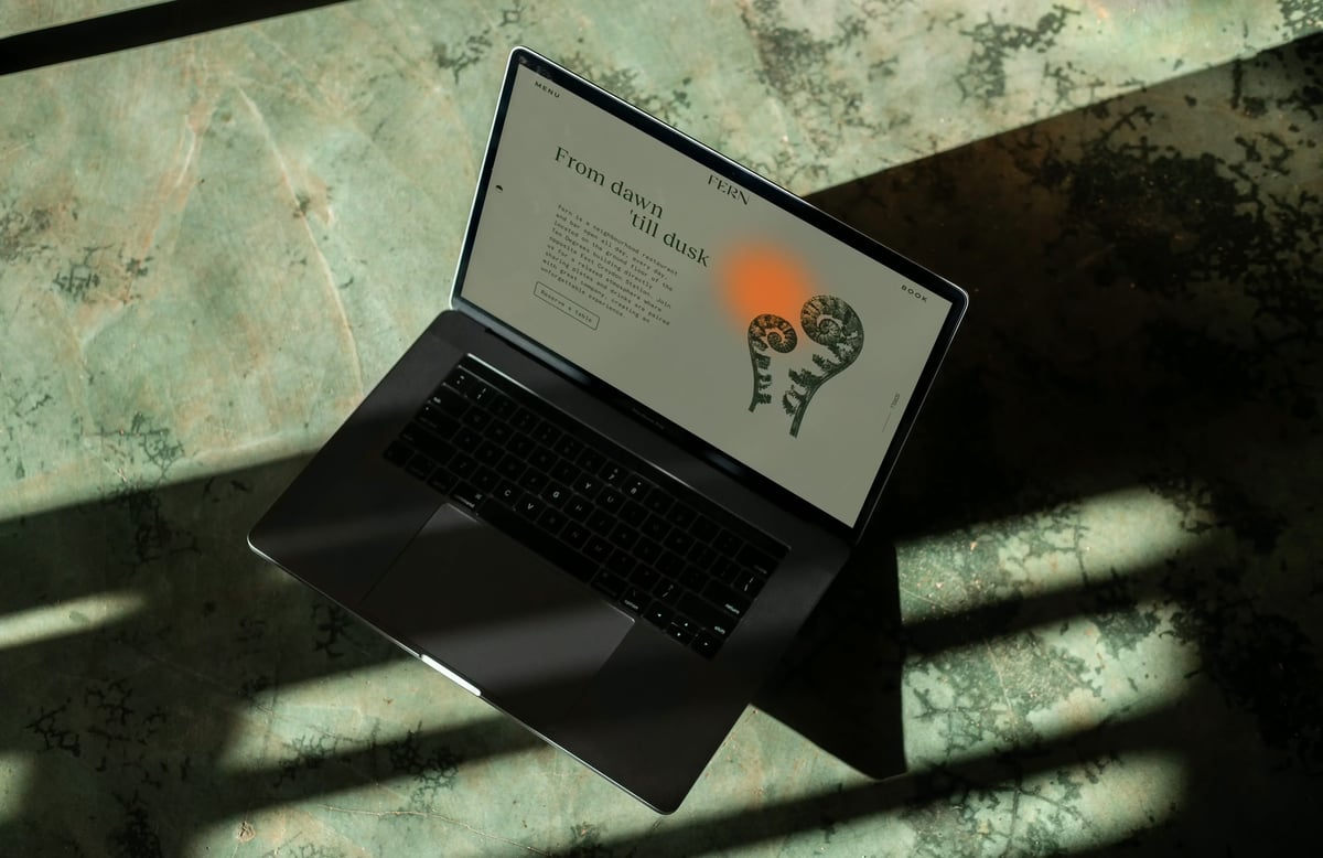 Laptop displaying the Fern website homepage with a sage green background, serif headline 'From dawn 'till dusk', a fern illustration, and a 'Reserve a Table' button.