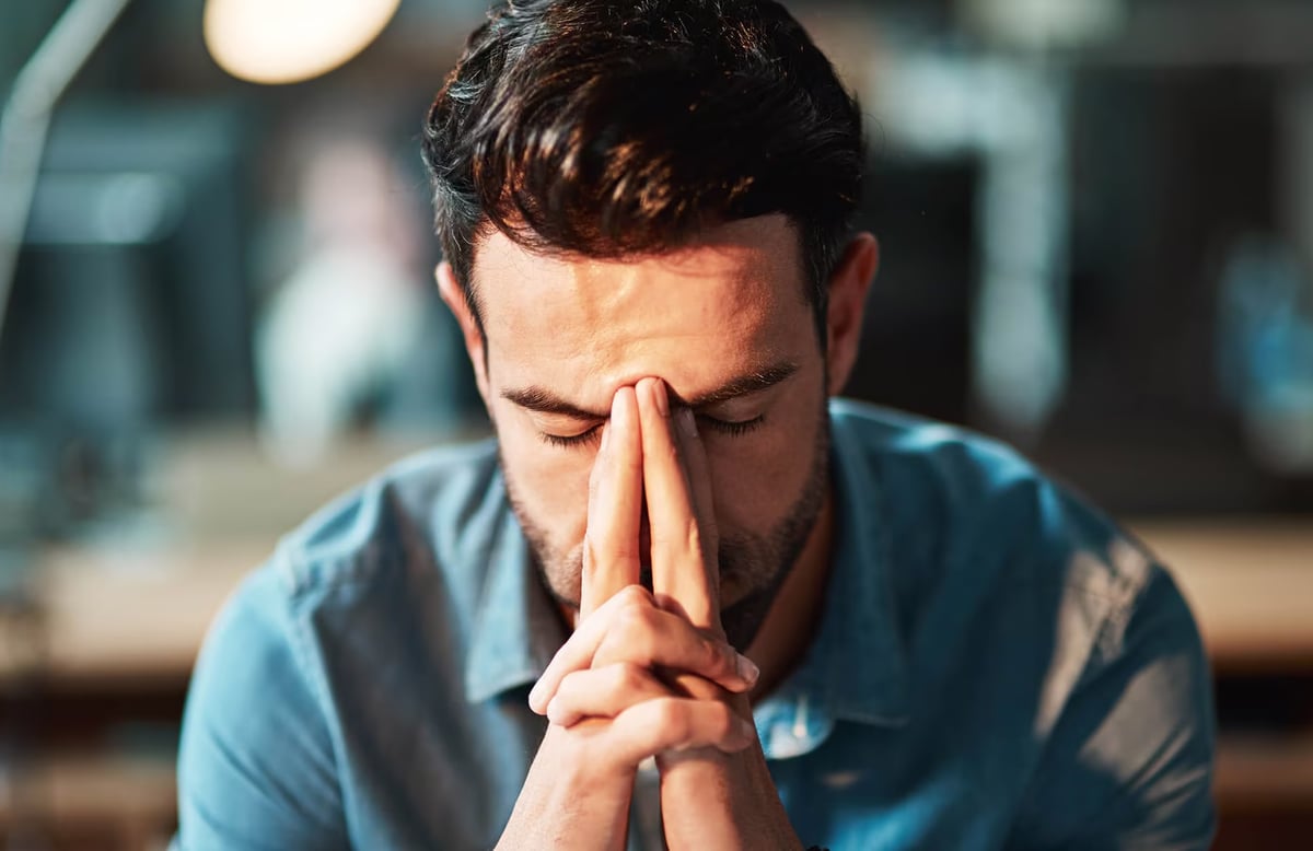 Close-up of a dark-haired man in a blue shirt sitting at a illuminated desk with his eyes closed, pressing his fingers to the bridge of his nose in frustration.