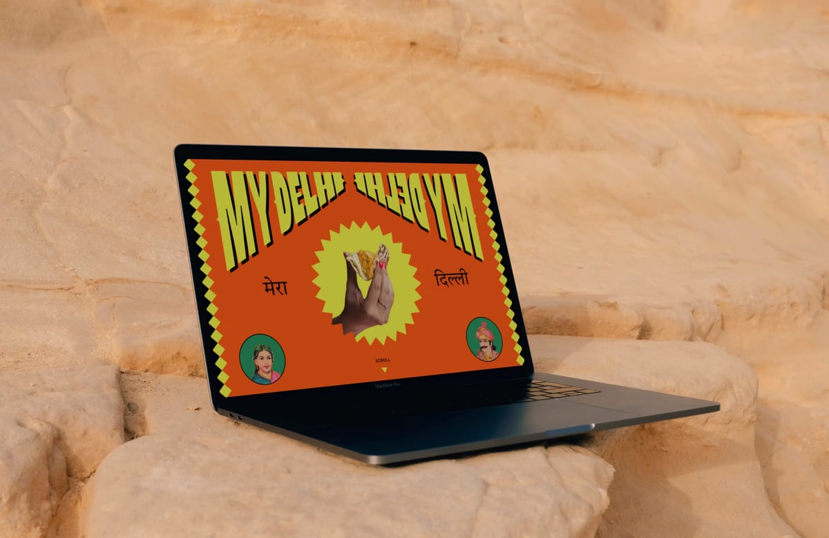 Laptop displaying the vibrant My Delhi website design featuring bold yellow arched typography, central food imagery inside a starburst, and Devanagari text on a bright orange background.