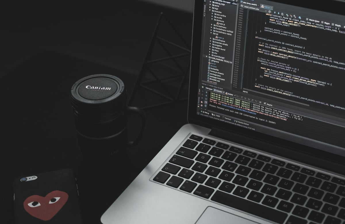 A dark minimalist desk setup featuring a laptop open to a code editor displaying JavaScript files next to a camera-lens mug and smartphone.
