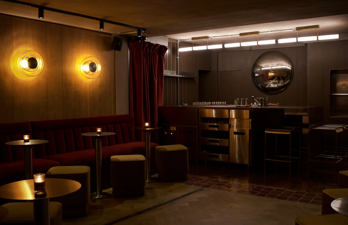 The moody, dimly lit basement lounge at Cato, featuring plush red velvet banquettes, warm circular sconces, candlelit bistro tables, and a modern stainless steel bar in the background.