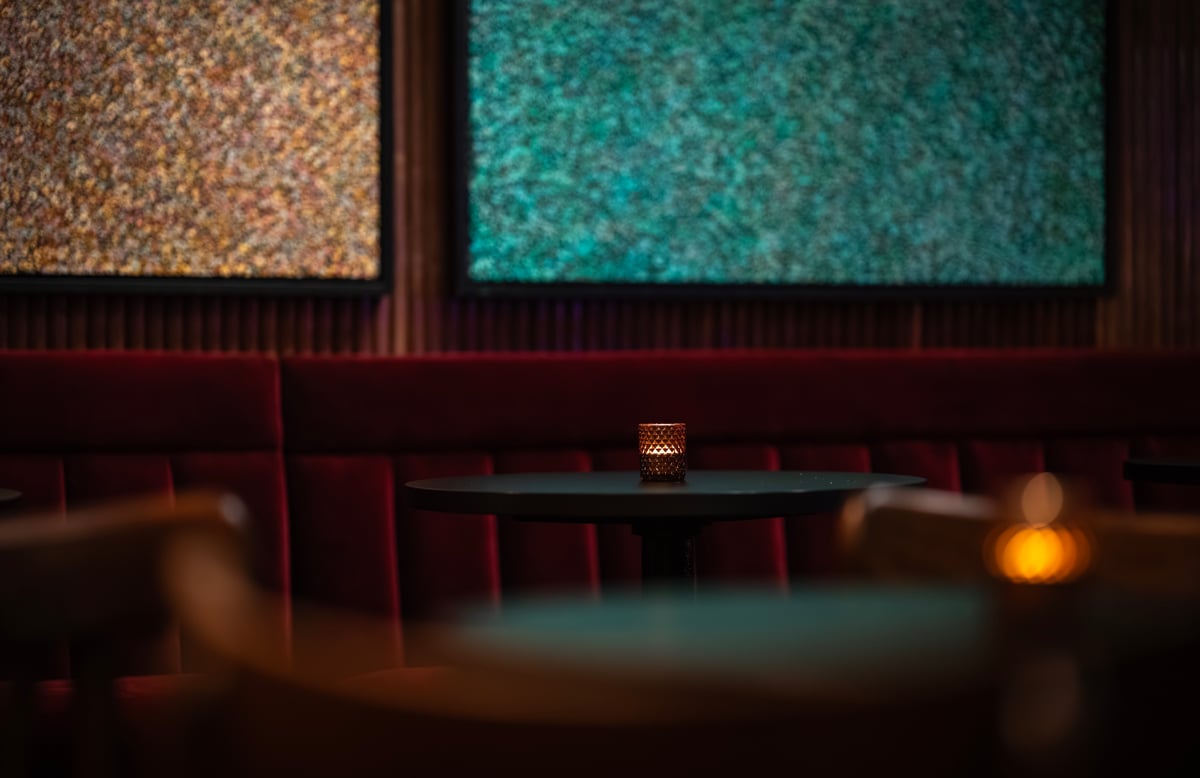 A lit candle on a dark round table inside the moody interior of The House of Julep, featuring plush red velvet seating and textured abstract wall art.