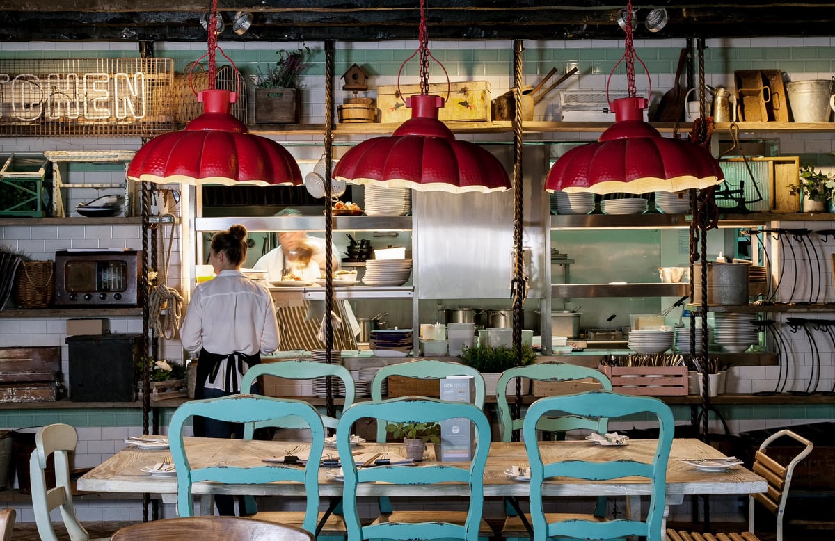 Rustic restaurant interior featuring turquoise dining chairs, wooden tables, and three prominent red pendant lights hanging in front of an open kitchen.