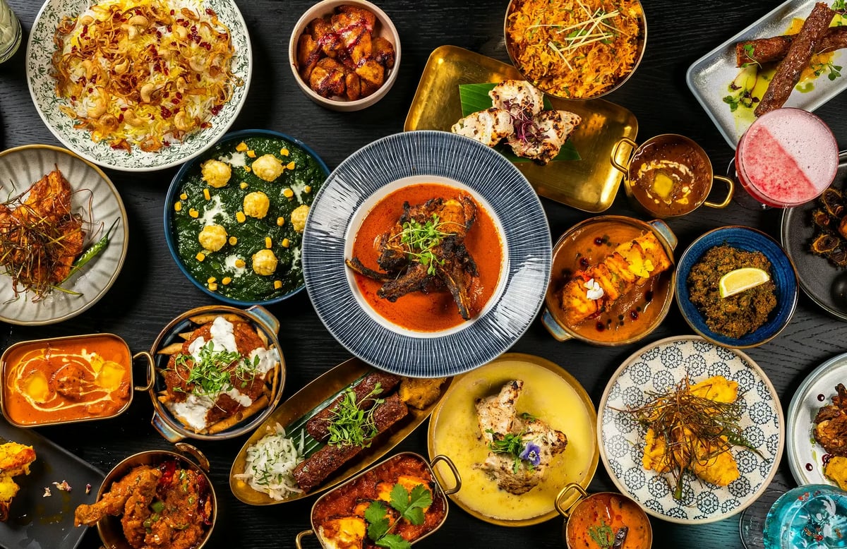 op-down flat lay view of a dark wooden table covered in a wide variety of colorful Indian dishes, curries, biryani, kebabs, and vibrant cocktails.