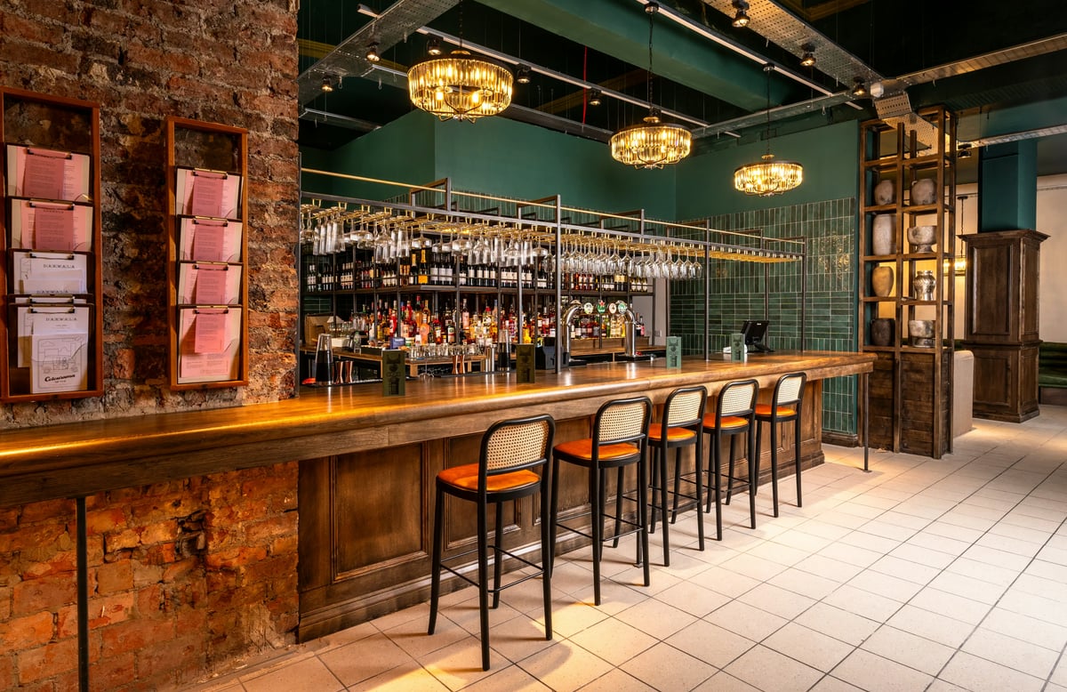 A stylish, dimly lit restaurant bar featuring a long wooden counter lined with high cane-back stools, hanging wine glasses, and warm chandelier lighting against teal walls and exposed brickwork.