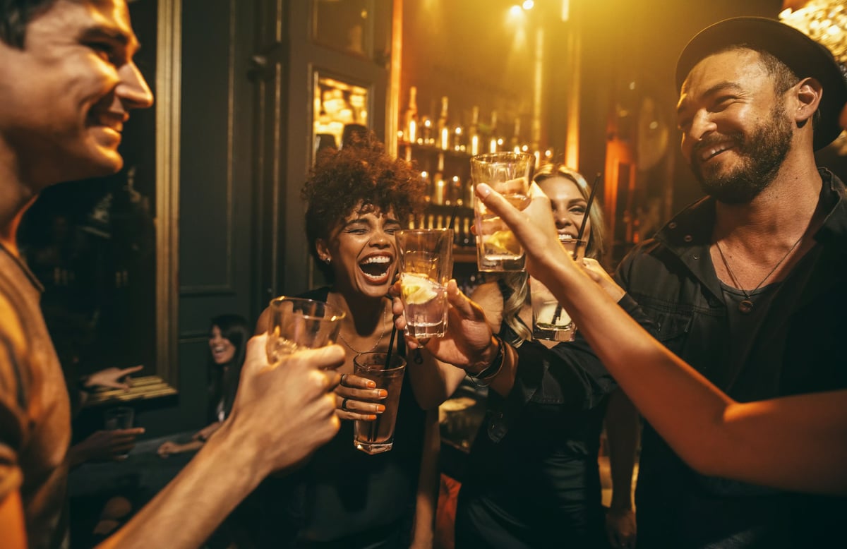 A diverse group of friends smiling and clinking cocktail glasses together at a warmly lit bar.