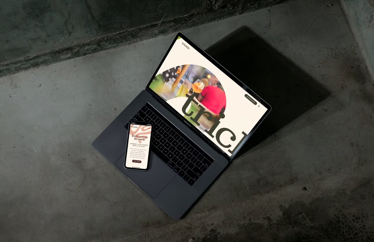 A laptop and overlapping smartphone displaying the desktop and mobile versions of the Trickle website, featuring bold typography, arched food imagery, and a 'Bottomless Brunch' promo screen.