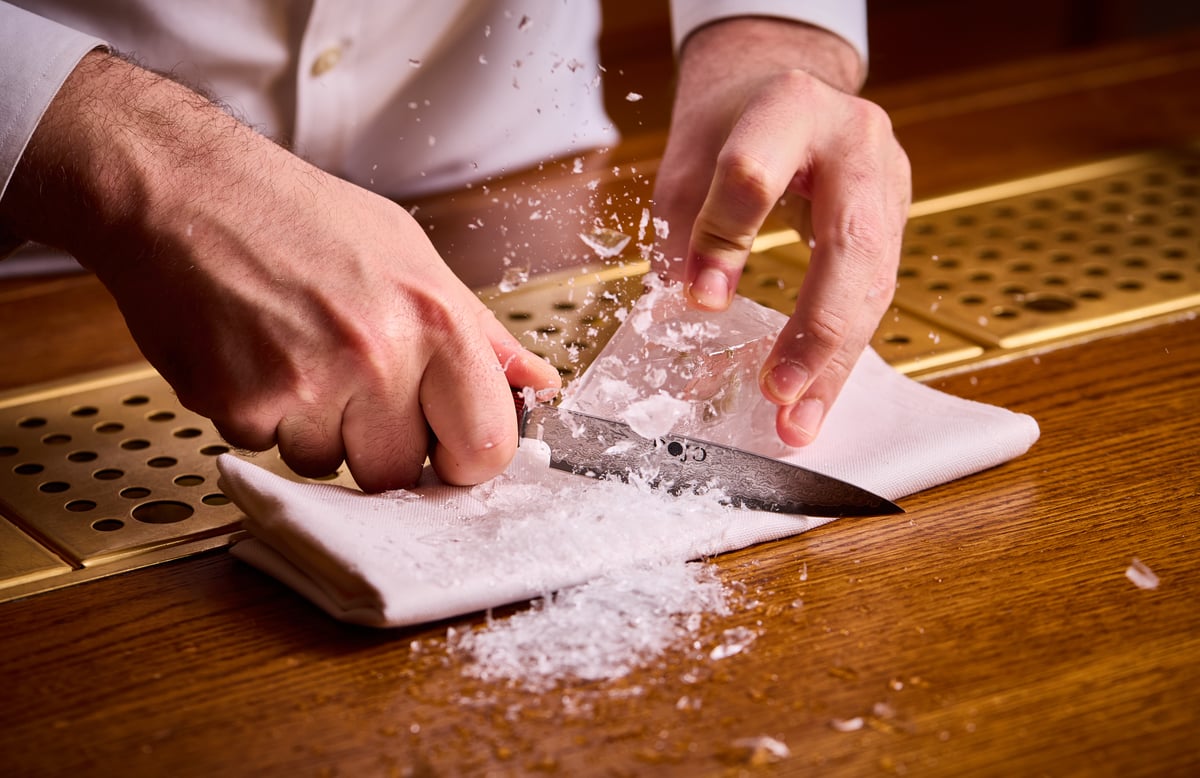 Close-up photograph of a bartender hand-carving a clear block of ice with a sharp knife on a folded white towel at a wooden bar, with ice shavings spraying into the air.