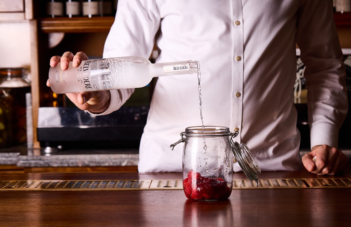 Action shot of a bartender pouring vodka over fresh raspberries in a glass jar.
